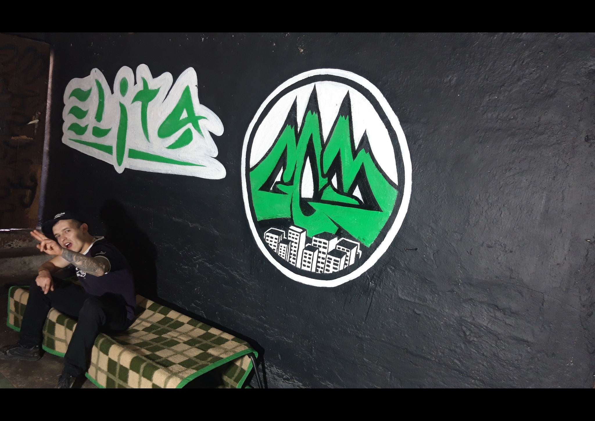 Młody mężczyzna siedzi na tapczanie na tle ściany z graffiti w odcieniach zieleni i bieli, przedstawiających napis 'Elita' i logo z motywem gór i miasta.