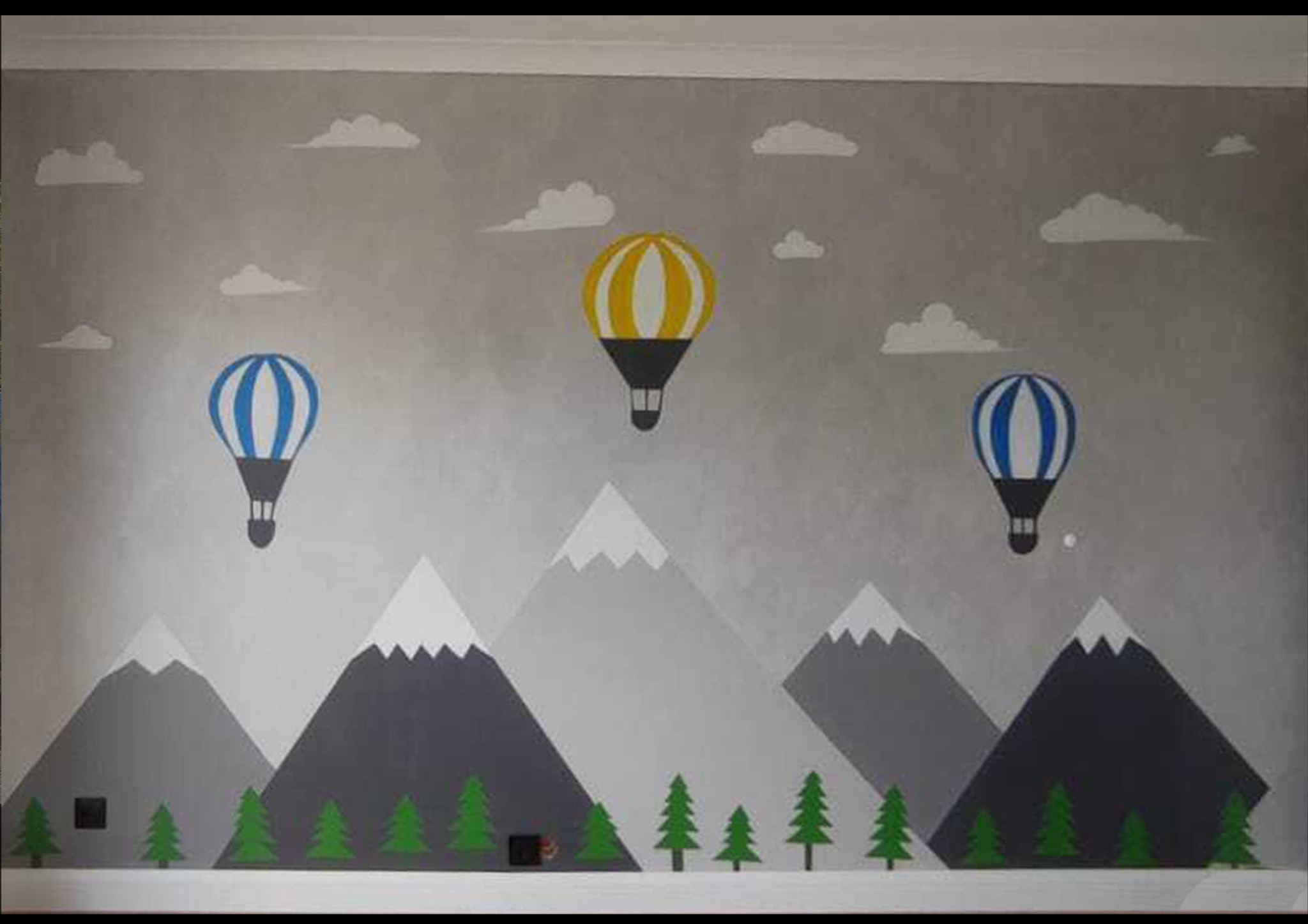 Pokój dziecięcy z ręcznie malowaną ścianą przedstawiającą górski krajobraz z balonami na tle chmur.