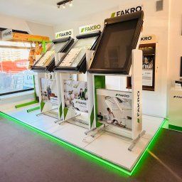 Vena Dach Katarzyna Miotke - Ekspozycja okien dachowych Fakro w salonie: trzy modele z roletami, każdy na białym stojaku z zielonymi akcentami i reklamami. Podświetlana podłoga LED.