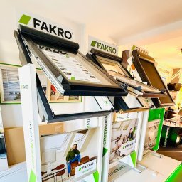 Vena Dach Katarzyna Miotke - Ekspozycja okien dachowych Fakro Greenview w salonie. Różne modele z roletami i bez, prezentowane na stojakach z grafikami. Widoczne żaluzje w tle.