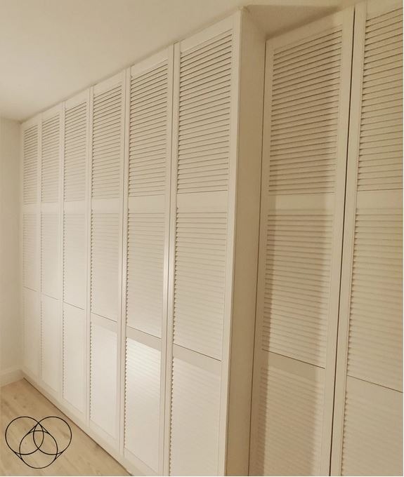 Długa, biała szafa wnękowa z żaluzjowymi drzwiami, wypełniająca całą ścianę od podłogi do sufitu, widoczna pod kątem.