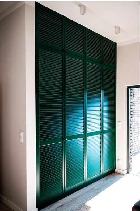 Wysoka, zabudowana szafa wnękowa z zielonymi, żaluzjowymi drzwiami, sięgająca sufitu, z małymi, srebrnymi uchwytami, w jasnym przedpokoju z szarą podłogą.