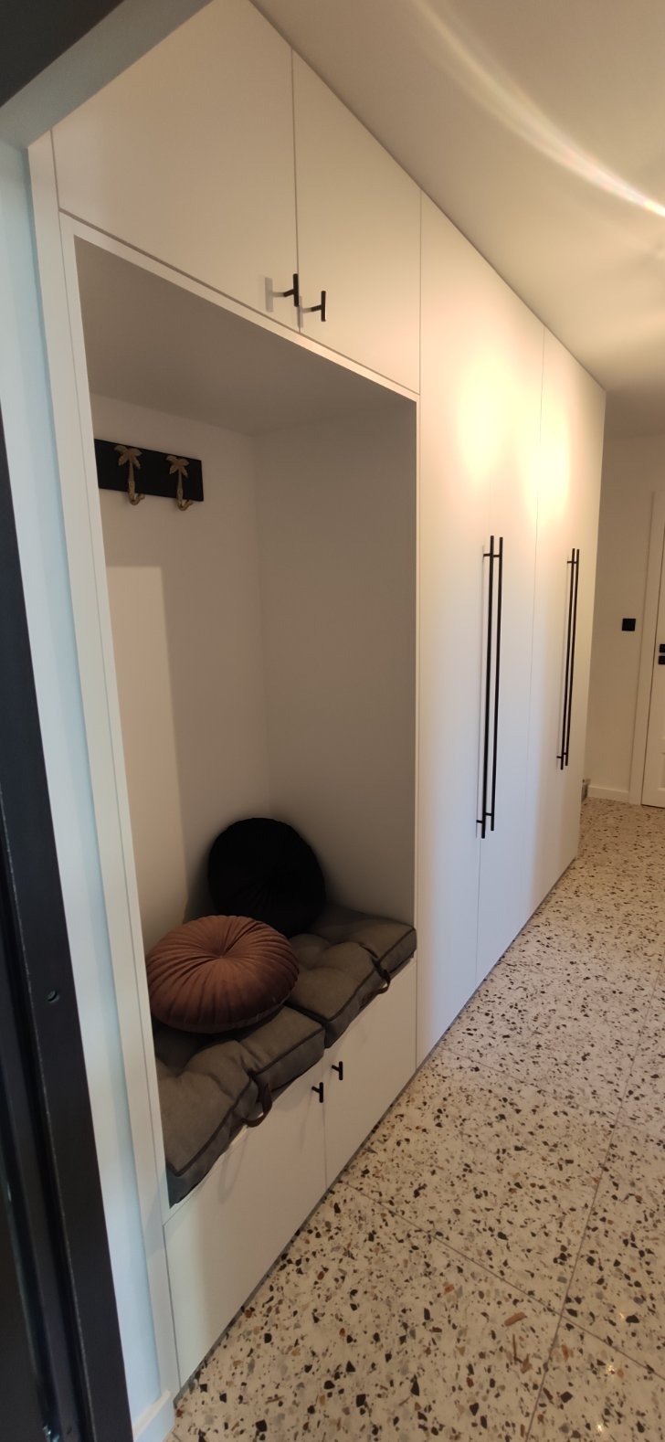 Wnęka z siedziskiem i poduszkami w zabudowie szafy w przedpokoju, z wieszakami na ścianie i terazzo na podłodze.
