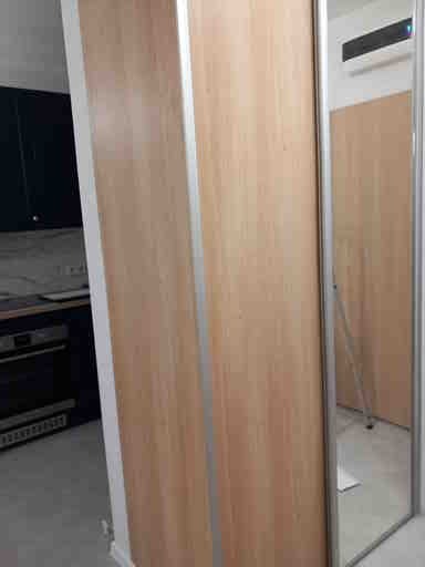 Jasna szafa wnękowa z przesuwnymi drzwiami z płyty imitującej drewno, z wąskimi srebrnymi listwami pionowymi i lustrzanym panelem po prawej stronie, w tle widoczna kuchnia.