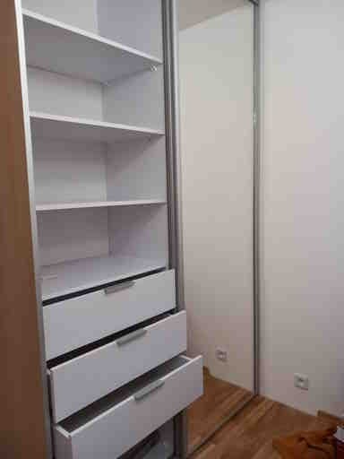 Wnętrze szafy z białymi półkami i szufladami z srebrnymi uchwytami, widoczne przesuwne drzwi z lustrem i fragment drewnianej podłogi.