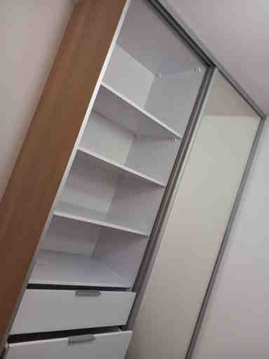 Wnętrze szafy z przesuwnymi drzwiami, widoczne półki i szuflady w kolorze białym, korpus w odcieniu drewna, z metalowymi prowadnicami.