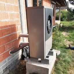 Centrum Rozwiązań Grzewczych KRAN - Zewnętrzna jednostka pompy ciepła zamontowana na betonowym fundamencie przy ścianie budynku z czerwonej cegły. Widoczne rury przyłączeniowe, fragment trawnika i narzędzie w tle.