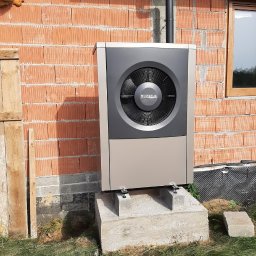 Centrum Rozwiązań Grzewczych KRAN - Zewnętrzna jednostka pompy ciepła Buderus zamontowana na betonowym fundamencie przy ścianie z czerwonej cegły w surowym stanie, widoczny fragment rynny i okna.