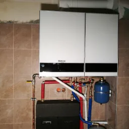 Biały, dwudrzwiowy piec grzewczy marki Buderus zamontowany na ścianie z brązowych płytek, z widoczną instalacją rur miedzianych, czerwonych i niebieskich, oraz dwoma zbiornikami wyrównawczymi...