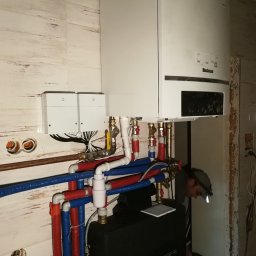 Centrum Rozwiązań Grzewczych KRAN - Instalacja kotła grzewczego Buderus z widocznymi rurami w izolacji niebieskiej i czerwonej, zaworami, oraz pracującym instalatorem z lampą czołową w pomieszczeniu wykończonym płytkami imitującymi...