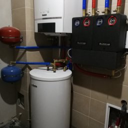 Centrum Rozwiązań Grzewczych KRAN - Kocioł gazowy Buderus z osprzętem: zbiornik, naczynia przeponowe, rury w izolacji niebieskiej i czerwonej, zamontowany na beżowych płytkach w pomieszczeniu technicznym.