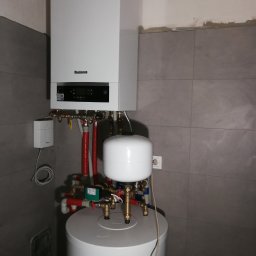 Centrum Rozwiązań Grzewczych KRAN - Biały, wiszący piec gazowy marki Buderus z podłączonym zasobnikiem ciepłej wody, widoczne rury i zawory, szare płytki na ścianie.