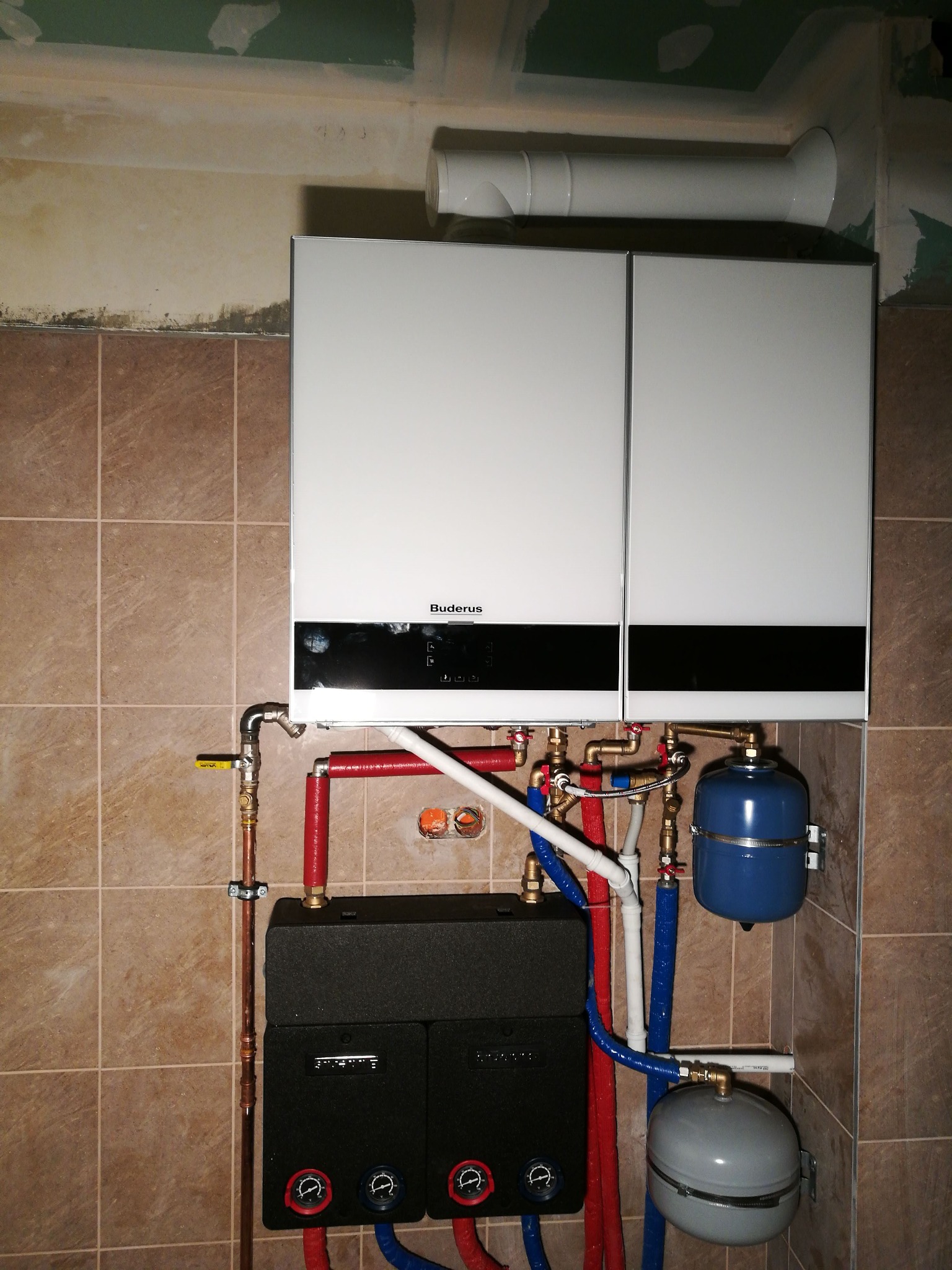 Biały, dwudrzwiowy piec grzewczy marki Buderus zamontowany na ścianie z brązowych płytek, z widoczną instalacją rur miedzianych, czerwonych i niebieskich, oraz dwoma zbiornikami wyrównawczymi...