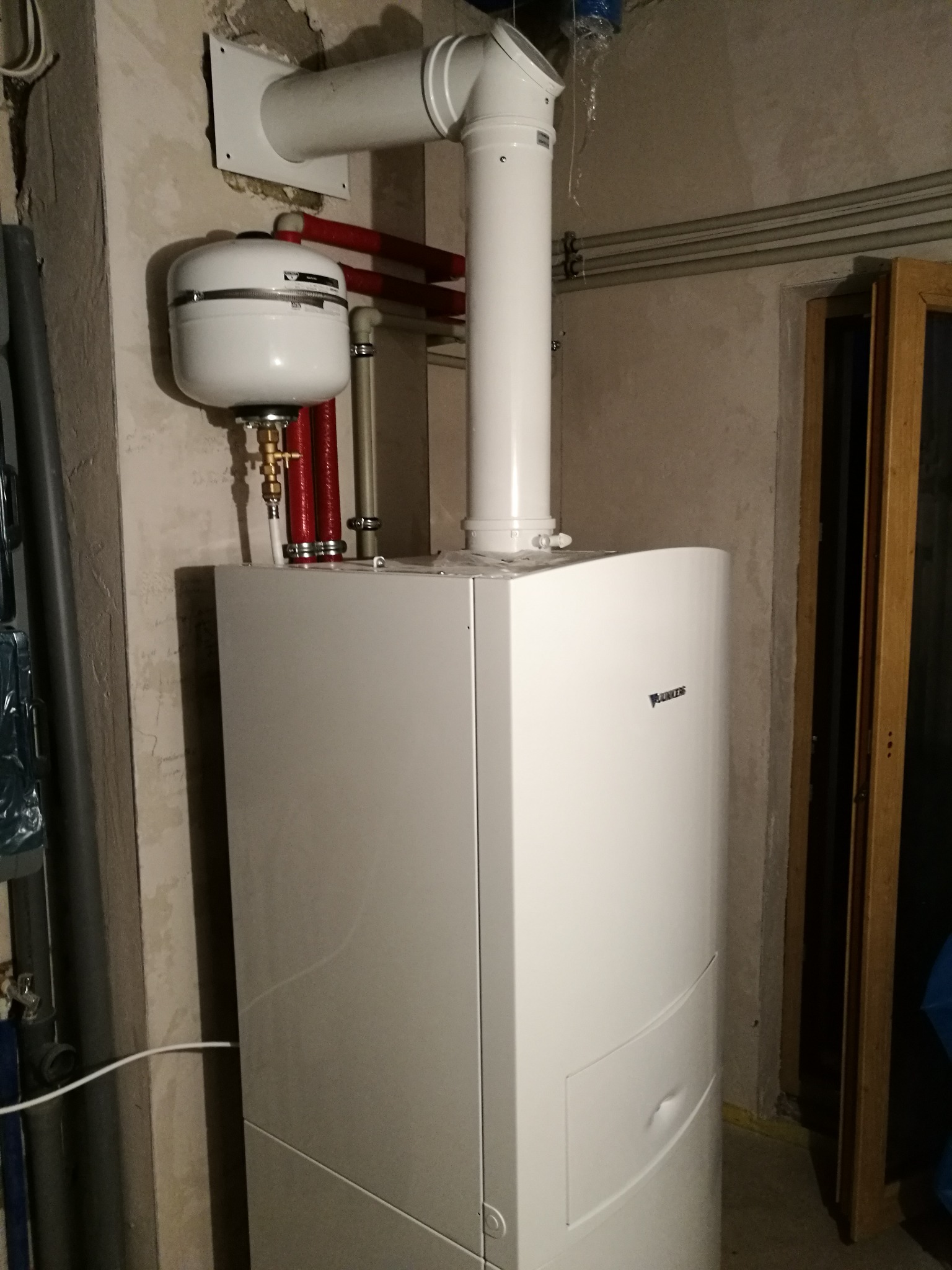 Biały piec grzewczy Daikin z podłączonymi rurami, zbiornik wyrównawczy i komin w surowym pomieszczeniu z widocznymi rurami na ścianie.