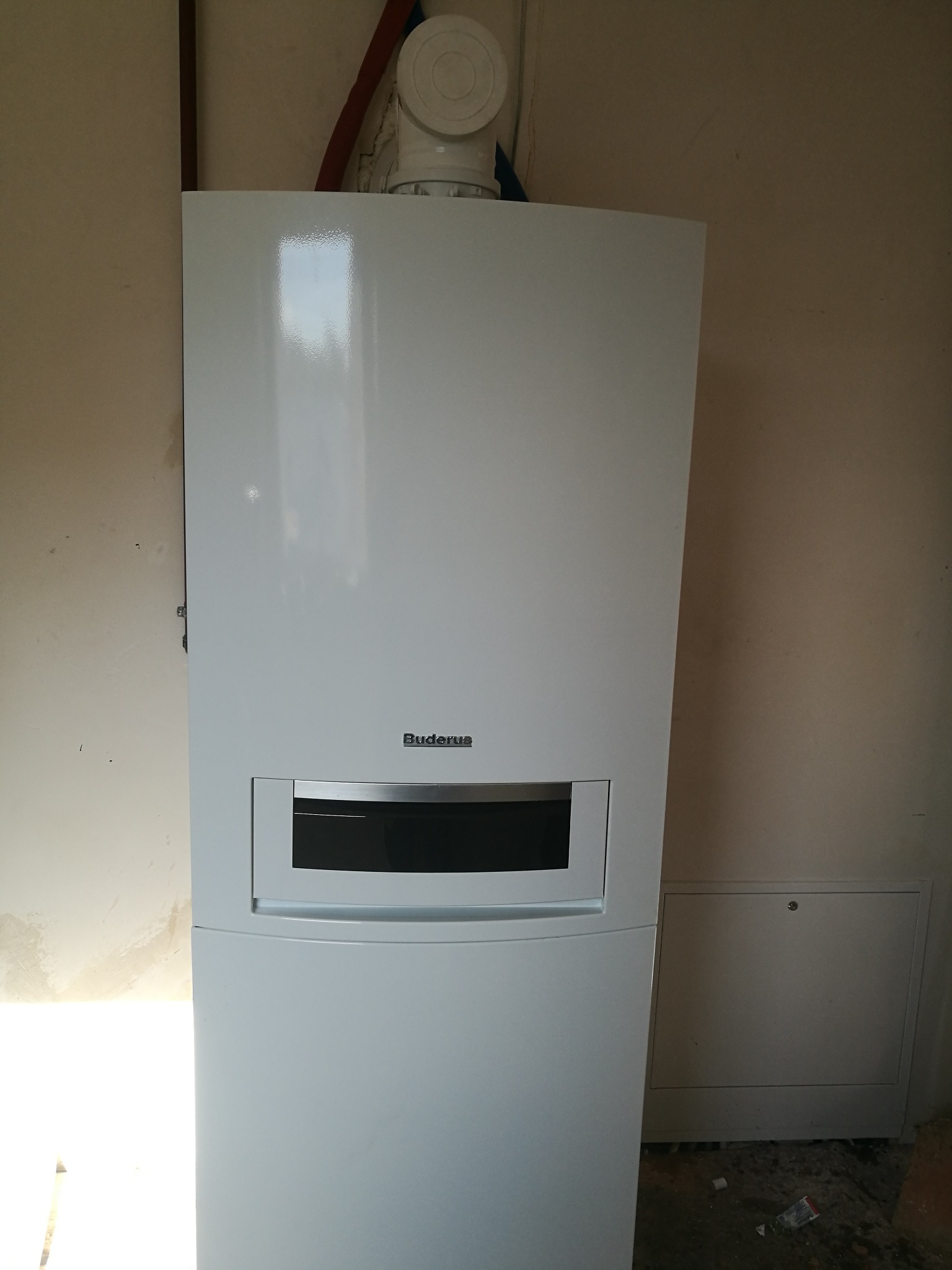 Biały, wiszący kocioł grzewczy marki Buderus zamontowany na ścianie w surowym wnętrzu. Widoczne rury instalacyjne i mała skrzynka rewizyjna obok kotła.