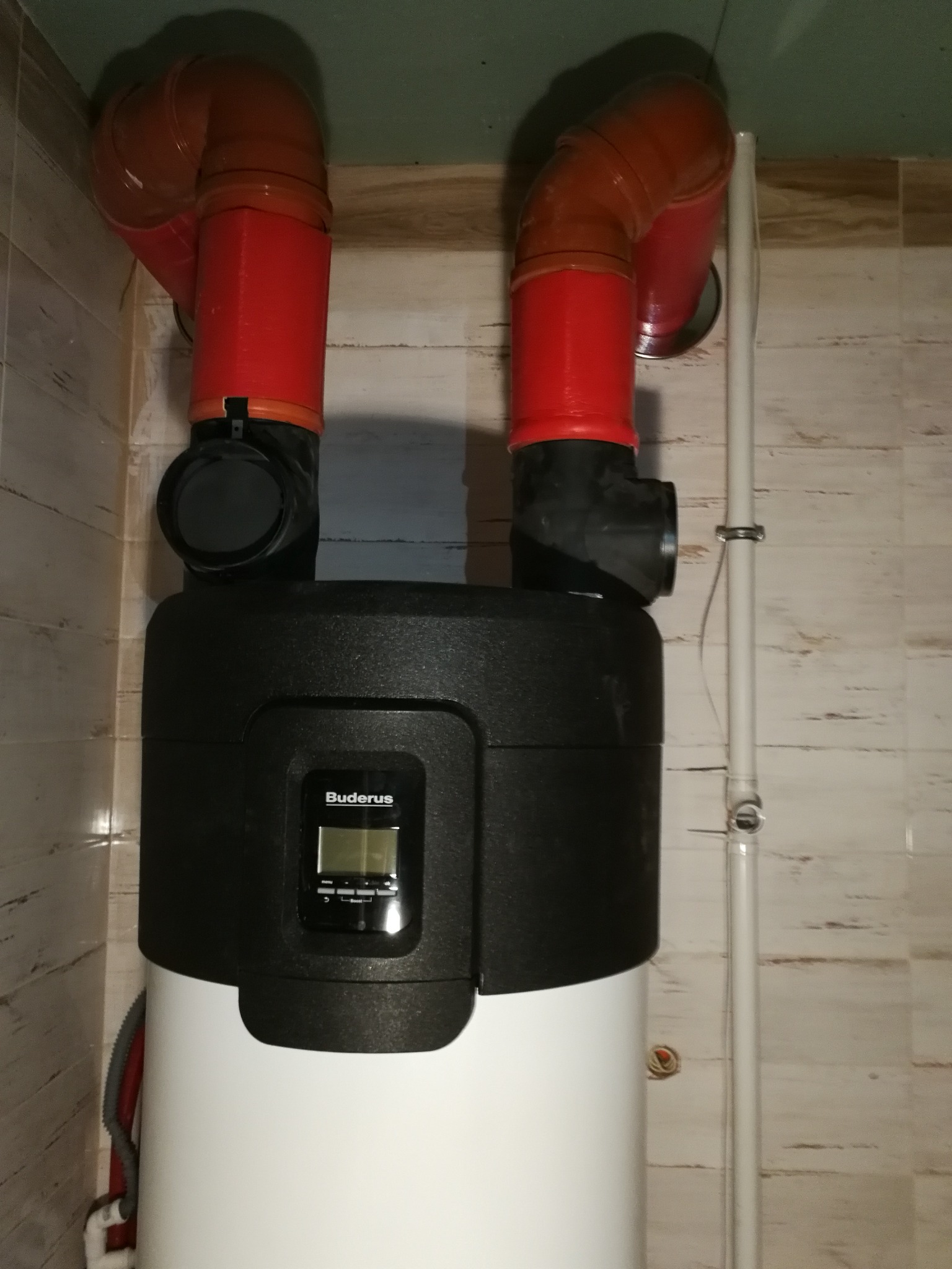 Biały bojler marki Buderus z czarną górną częścią i dwoma czerwonymi rurami odprowadzającymi spaliny, zamontowany w pomieszczeniu z wykończeniem imitującym drewno.