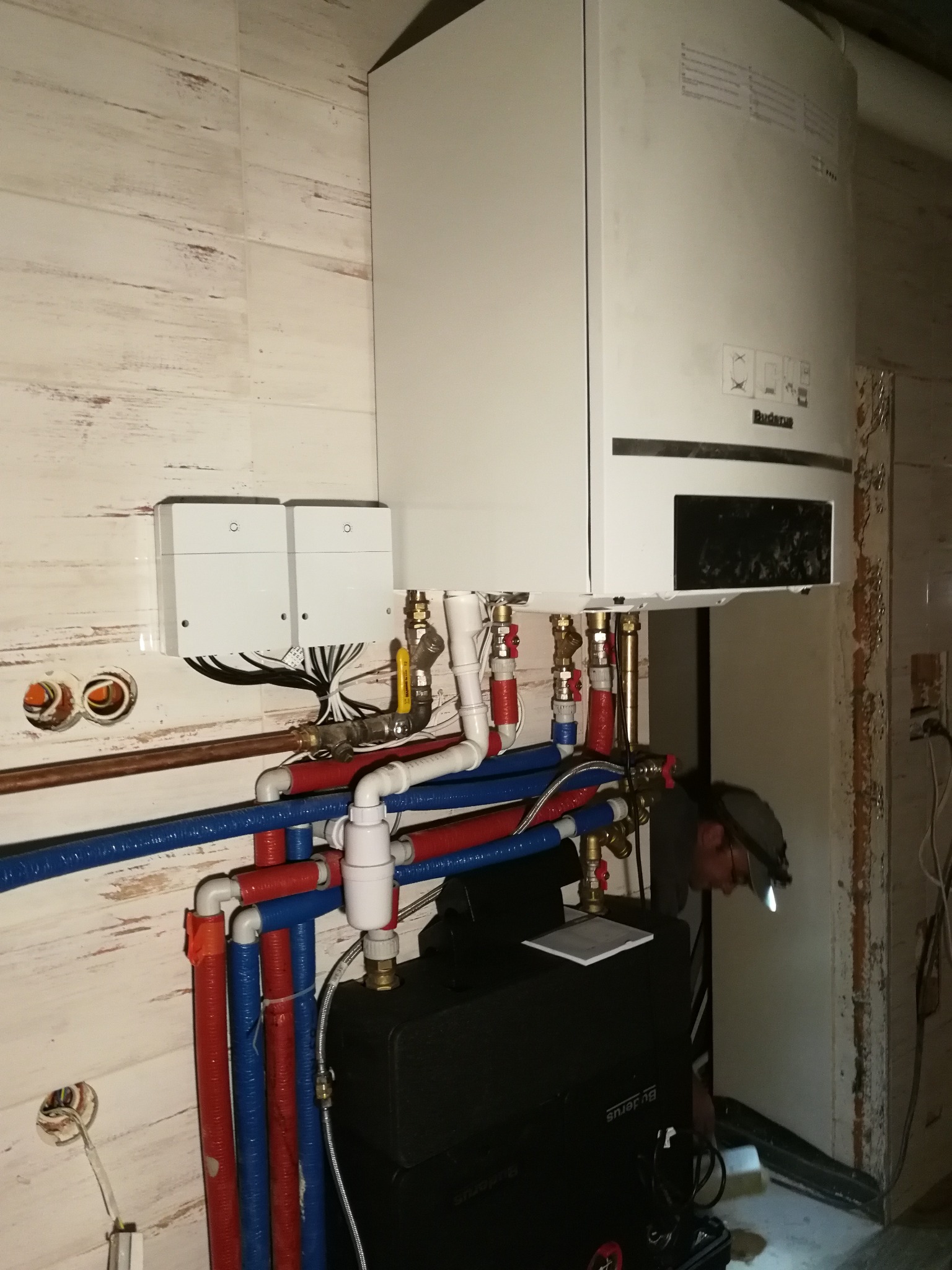 Instalacja kotła grzewczego Buderus z widocznymi rurami w izolacji niebieskiej i czerwonej, zaworami, oraz pracującym instalatorem z lampą czołową w pomieszczeniu wykończonym płytkami imitującymi...