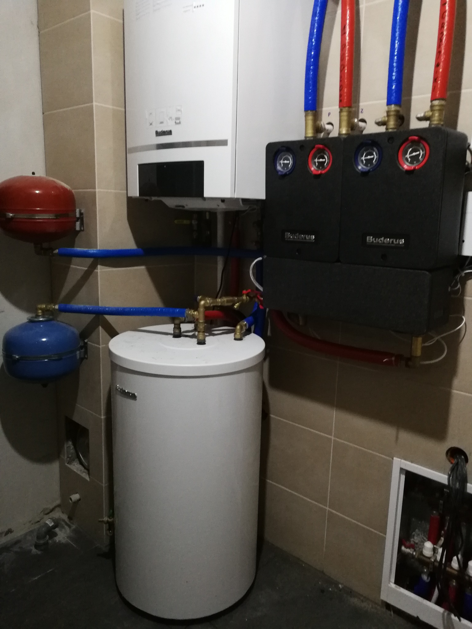 Kocioł gazowy Buderus z osprzętem: zbiornik, naczynia przeponowe, rury w izolacji niebieskiej i czerwonej, zamontowany na beżowych płytkach w pomieszczeniu technicznym.