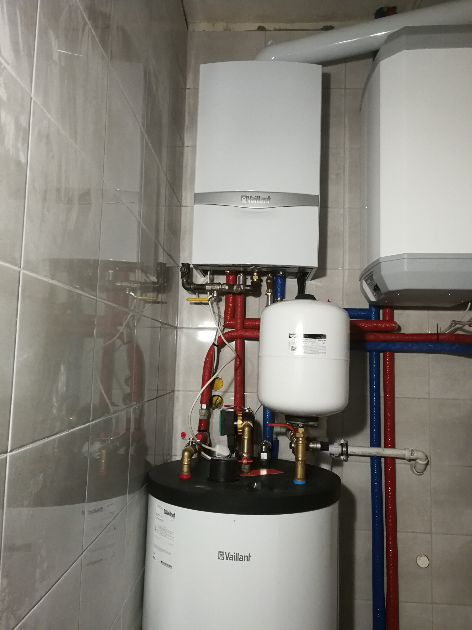 Kompaktowa kotłownia z wiszącym kotłem gazowym Vaillant, zbiornikiem ciepłej wody i naczyniem przeponowym, instalacja z rur miedzianych i tworzyw sztucznych, tło stanowią szare płytki ceramiczne.