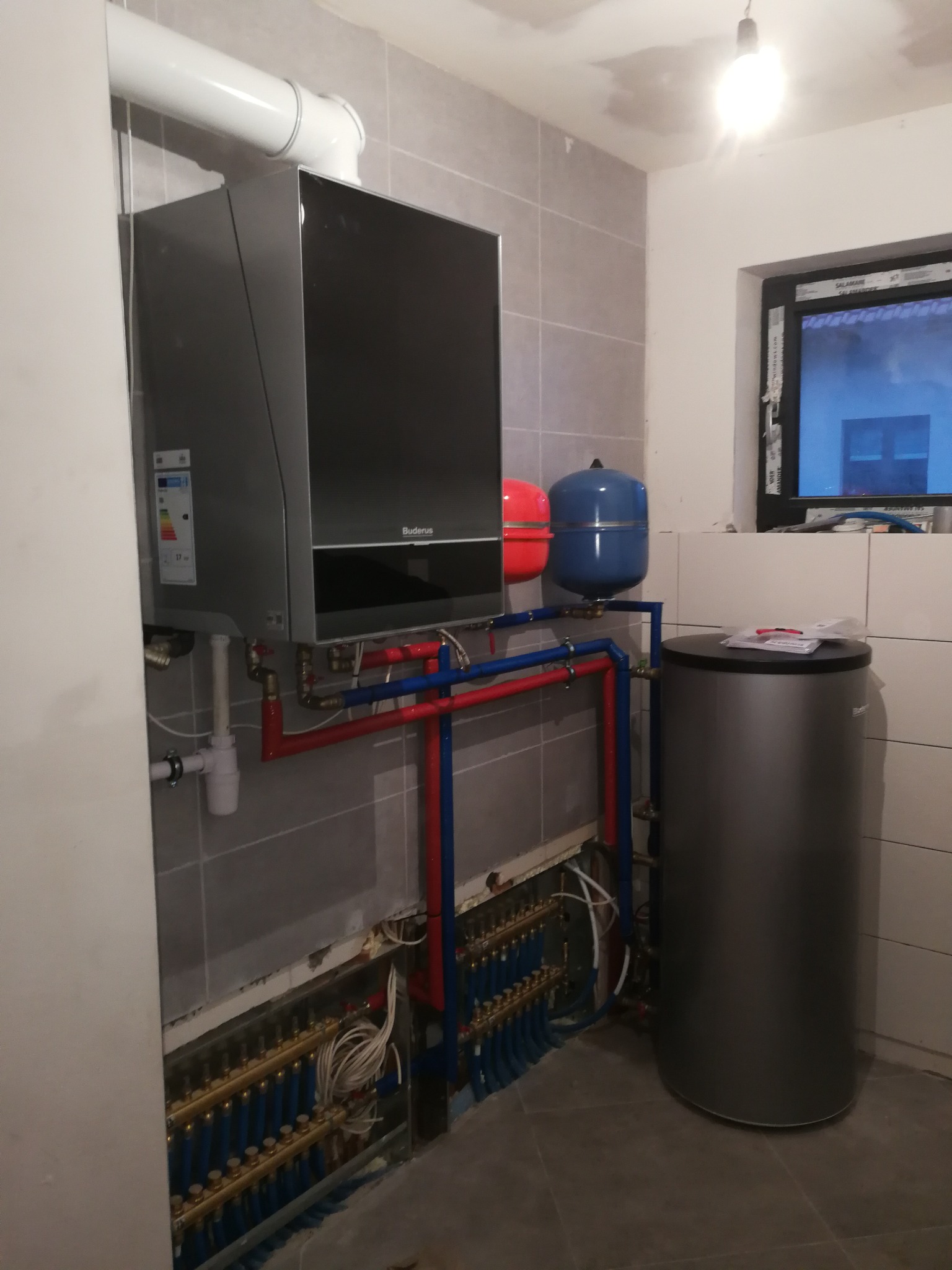 Szary kocioł grzewczy Buderus zawieszony na ścianie z szarych płytek, z widocznymi rurami w kolorze czerwonym i niebieskim, podłączony do rozdzielacza ogrzewania podłogowego i zbiornika na wodę,...