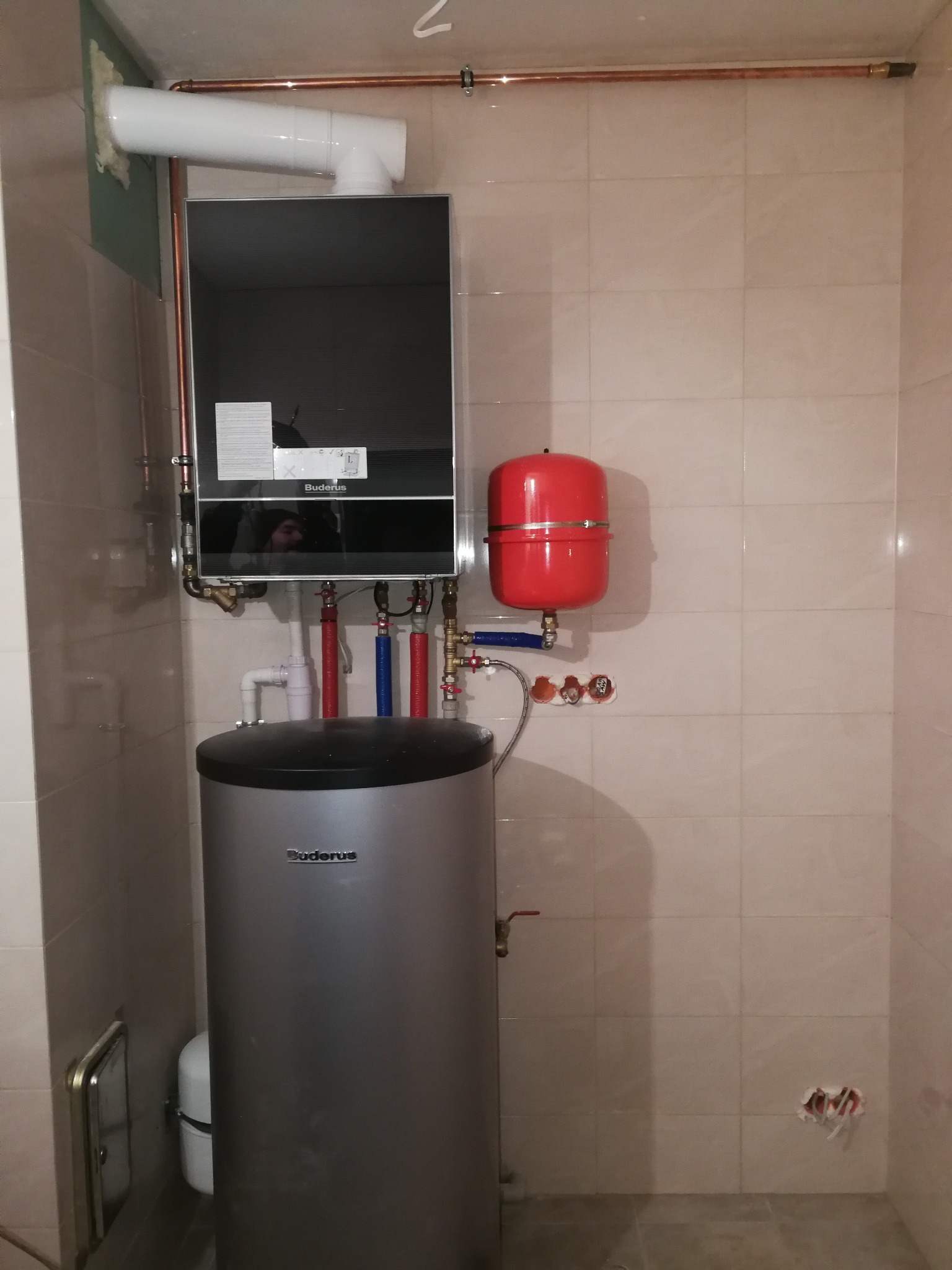 Zamontowany, wiszący, kondensacyjny kocioł gazowy Buderus z podłączonym zasobnikiem wody, rurami miedzianymi i czerwonym naczyniem przeponowym w łazience wyłożonej jasnymi płytkami.