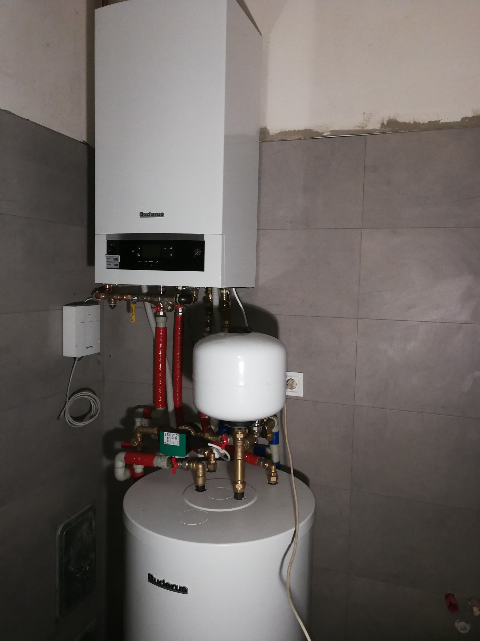 Biały, wiszący piec gazowy marki Buderus z podłączonym zasobnikiem ciepłej wody, widoczne rury i zawory, szare płytki na ścianie.