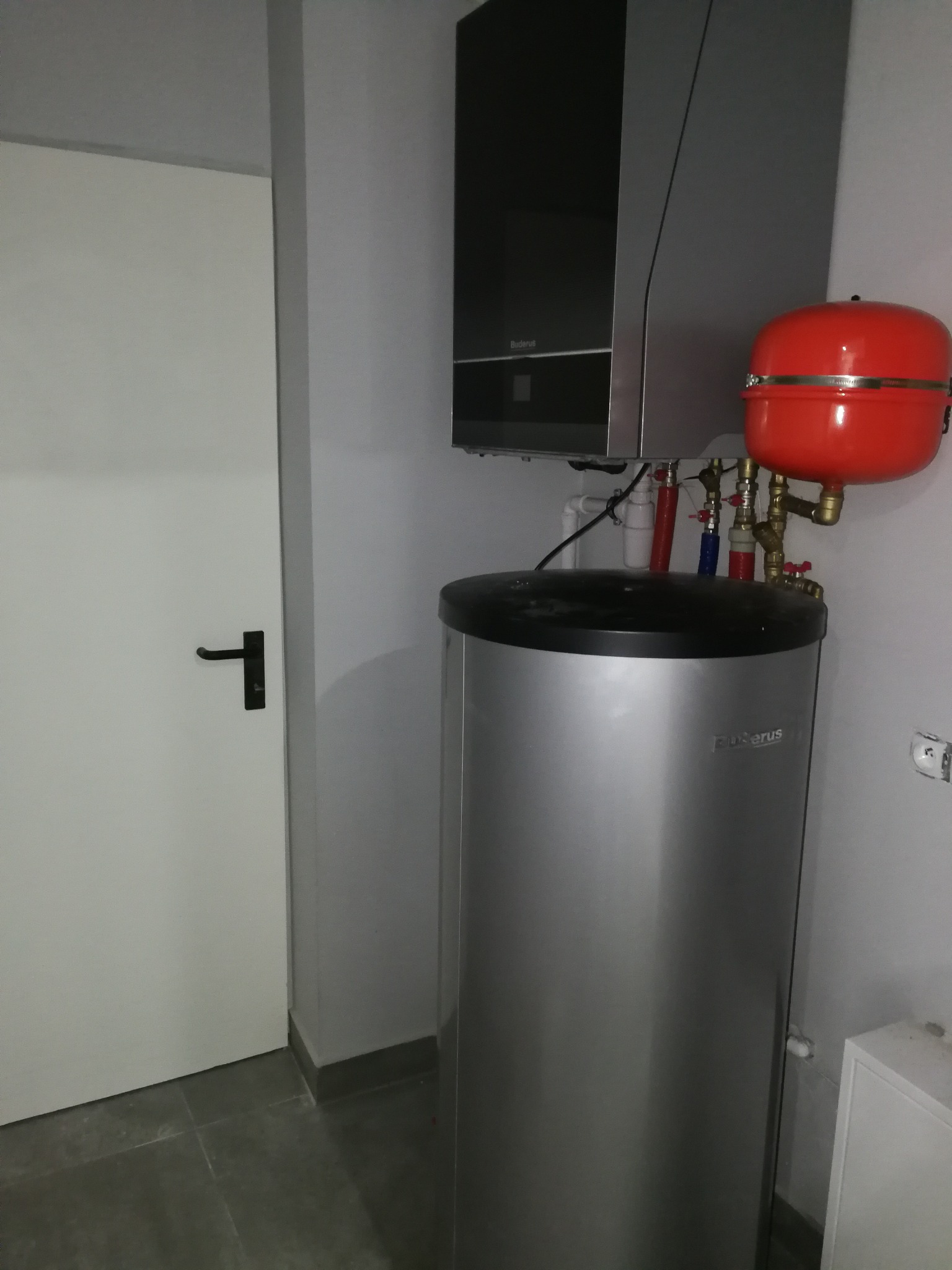 Kompaktowy zestaw grzewczy Buderus z czerwonym zbiornikiem wyrównawczym w kotłowni przy białych drzwiach z czarną klamką.