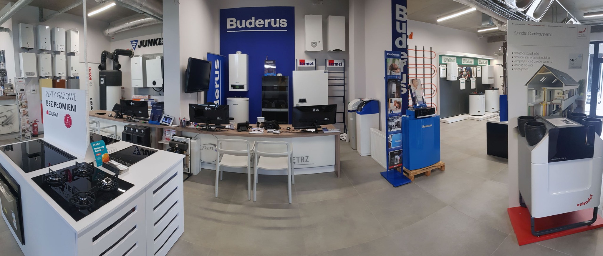 Ekspozycja w salonie z urządzeniami grzewczymi i instalacyjnymi różnych marek, w tym kotły gazowe, podgrzewacze wody, płyty gazowe bez płomieni, systemy wentylacyjne i rekuperacyjne; widoczny...