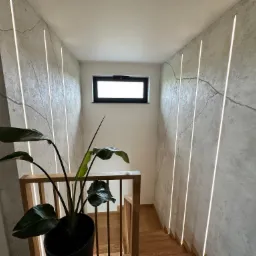 Schody z drewnianymi stopniami i balustradą, oświetlone pionowymi liniami LED na ścianach o fakturze betonu, z rośliną doniczkową w rogu i małym oknem na górze.