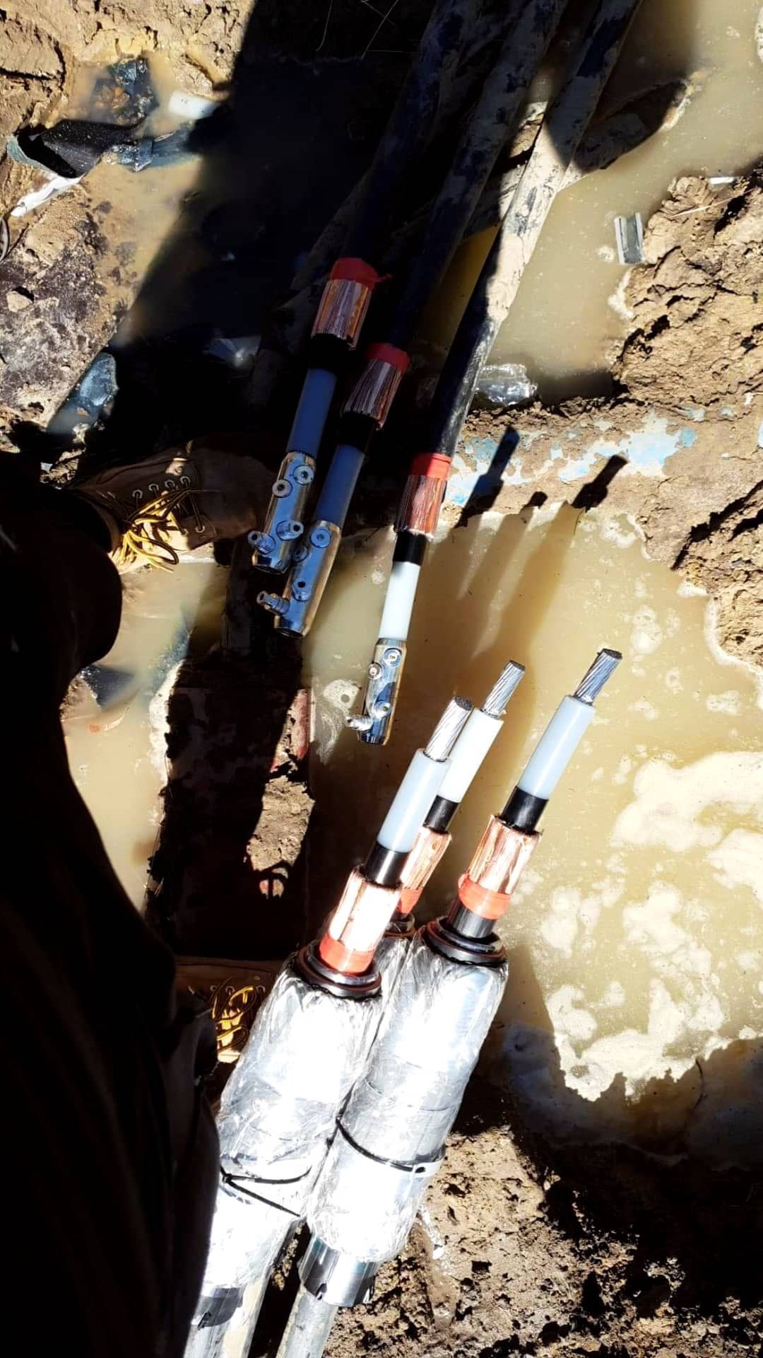 Podłączenie grubych kabli elektrycznych w wykopie wypełnionym wodą, widoczne końcówki kabli zabezpieczone taśmą i folią, elementy złączek i fragment buta roboczego.