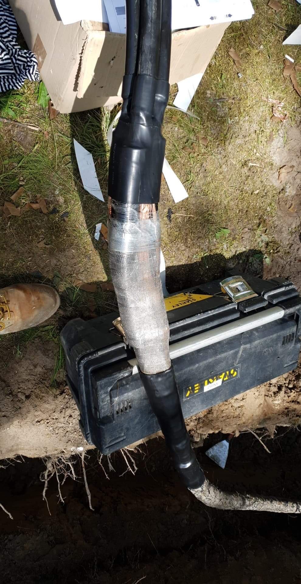 Połączenie kabli elektrycznych w ziemi, zabezpieczone taśmą izolacyjną i siatką ochronną, obok skrzynka narzędziowa Stanley i fragment buta roboczego