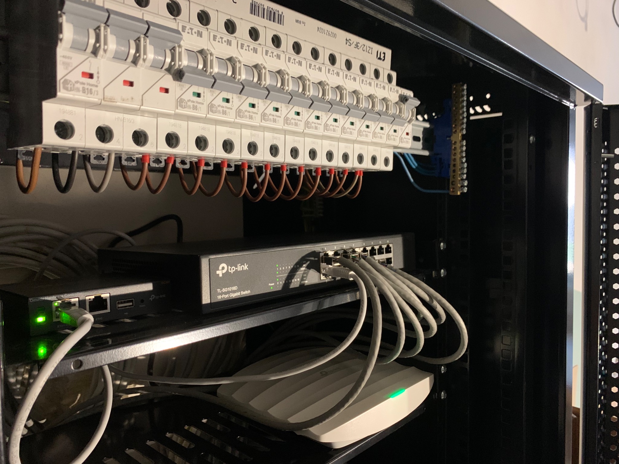 Szafa rack z zamontowanymi urządzeniami sieciowymi: switch TP-Link, router oraz punkt dostępowy Wi-Fi, powyżej listwa z bezpiecznikami Eaton z podłączonymi przewodami.