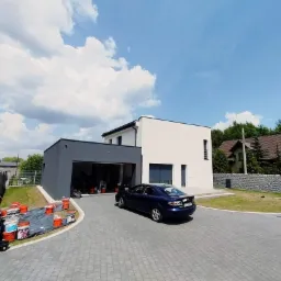 Nowoczesny dom jednorodzinny z szarą bryłą garażu, po świeżym pomalowaniu elewacji, widoczne wiaderka z farbą i folia ochronna na podjeździe z kostki brukowej.