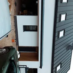 Nowoczesny dom jednorodzinny z białą elewacją i grafitowym dachem z blachy na rąbek stojący, widok z boku na garaż i podjazd z zaparkowanym samochodem.