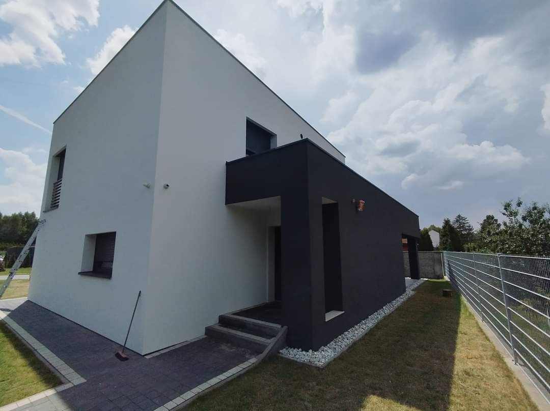 Dwukolorowy, nowoczesny dom jednorodzinny po odnowieniu elewacji, z białą częścią i antracytową bryłą, widoczna drabina oparta o ścianę, chodnik z kostki brukowej i trawnik.