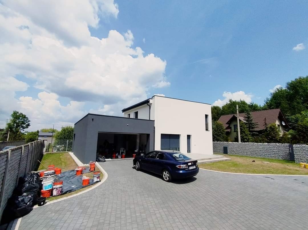 Nowoczesny dom jednorodzinny z szarą bryłą garażu, po świeżym pomalowaniu elewacji, widoczne wiaderka z farbą i folia ochronna na podjeździe z kostki brukowej.