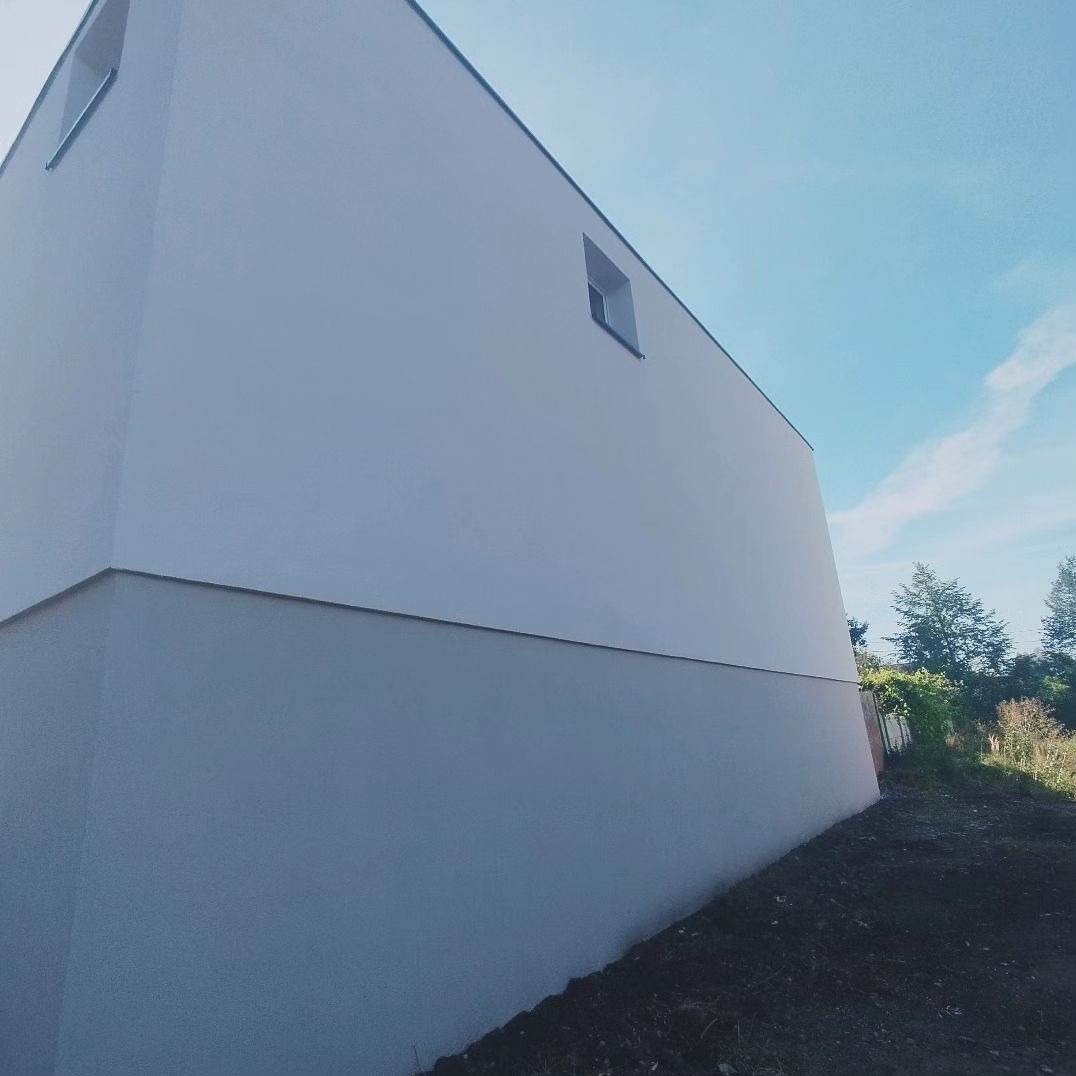 Minimalistyczny budynek z dwukolorową elewacją, górna część biała, dolna szara, widok z dołu, błękitne niebo w tle.
