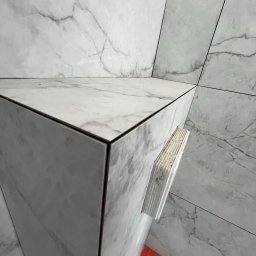 APPLI INVEST Mateusz Pieczarka - Wykończona narożna półka z płytek imitujących marmur, z widocznymi krawędziami i fugami. W tle ściana wyłożona tymi samymi płytkami. Widoczne elementy montażowe.