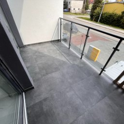 APPLI INVEST Mateusz Pieczarka - Wykończony balkon z szarymi płytkami i szklaną balustradą w Łężycy. Widok na ulicę. Nowoczesny design, staranne ułożenie płytek, czysty i estetyczny wygląd.