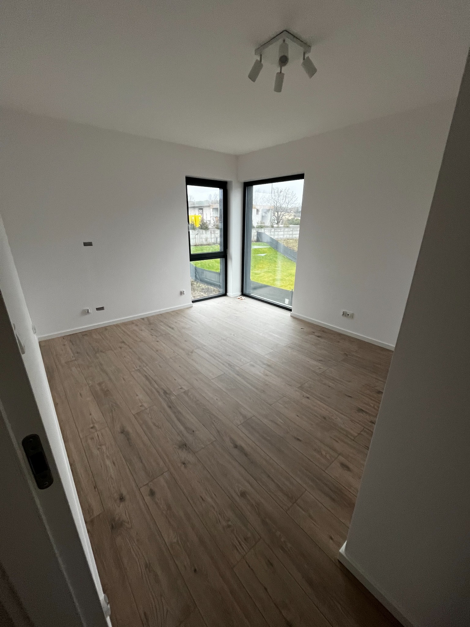 Jasne wnętrze z drewnopodobną podłogą i dużymi oknami narożnymi, przez które widać zielony trawnik. Minimalistyczne oświetlenie sufitowe.