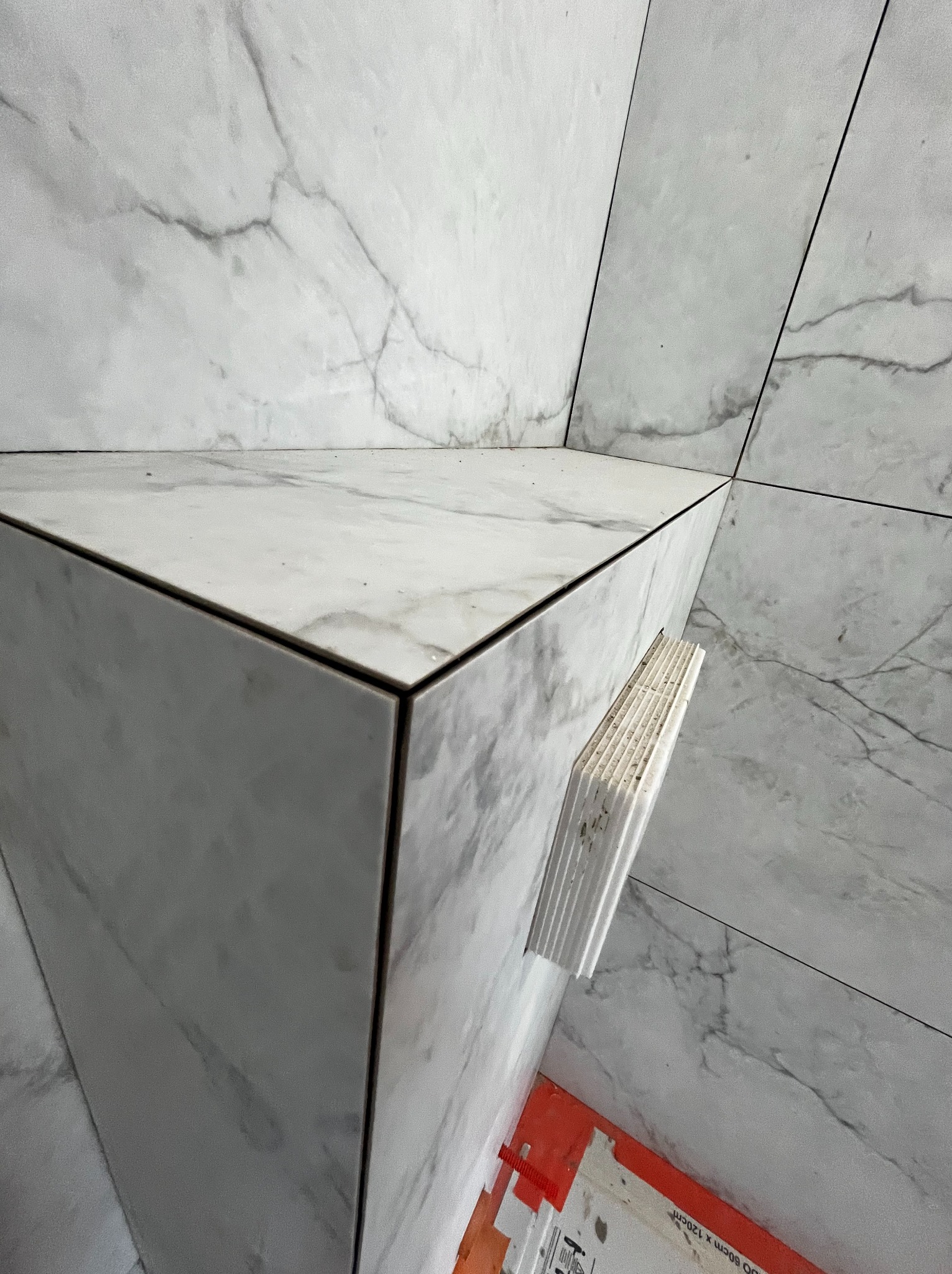 Wykończona narożna półka z płytek imitujących marmur, z widocznymi krawędziami i fugami. W tle ściana wyłożona tymi samymi płytkami. Widoczne elementy montażowe.