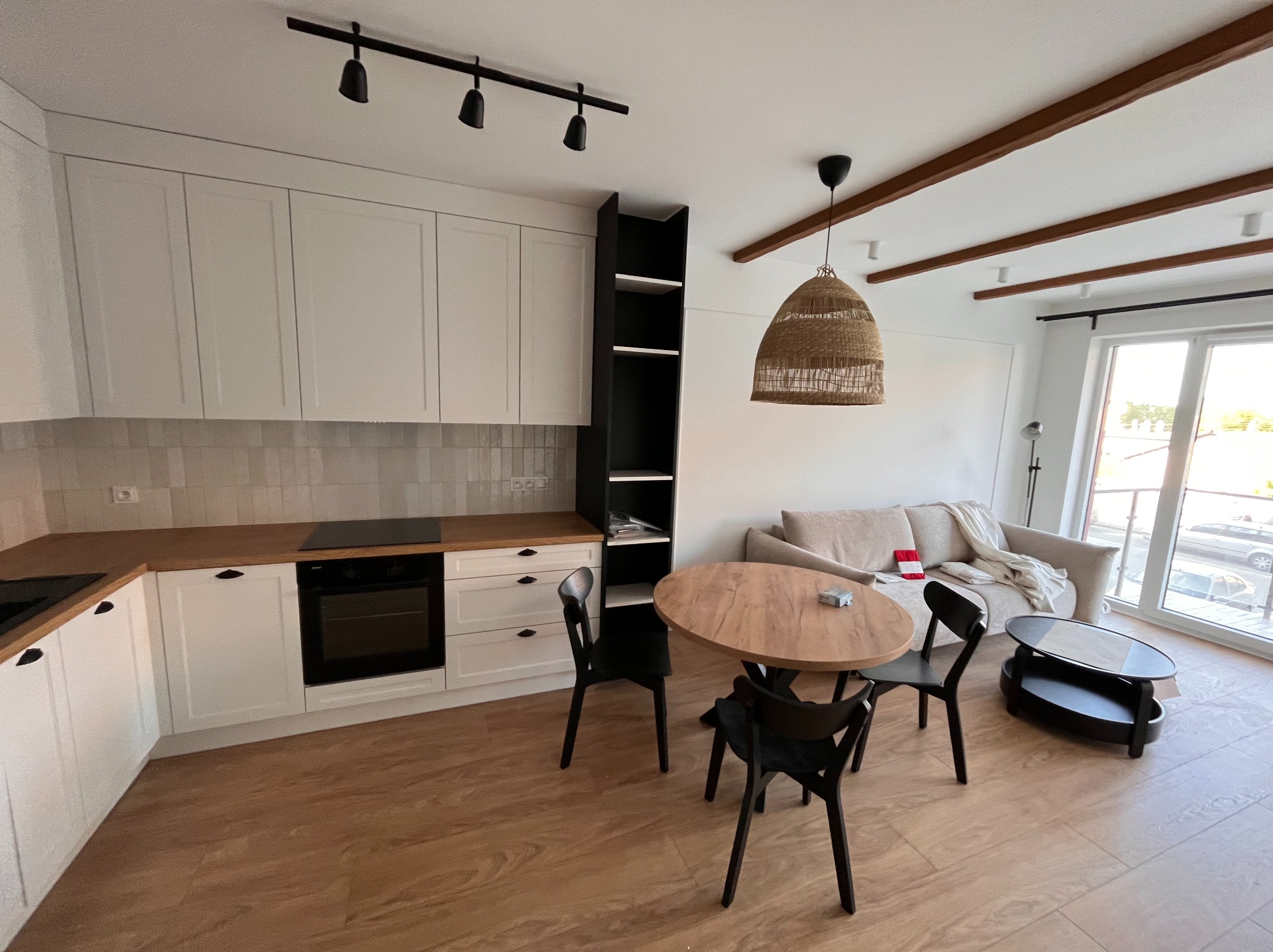 Jasna kuchnia z drewnianym blatem i płytkami, czarne krzesła przy okrągłym stole. W tle sofa i balkon. Nowoczesne oświetlenie sufitowe.