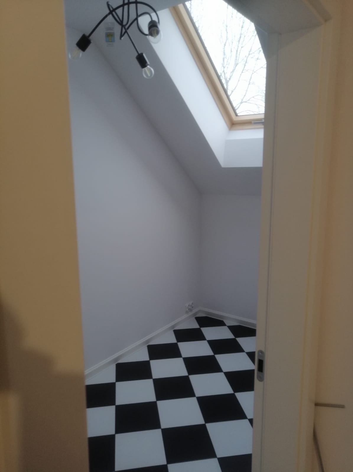 Wnętrze poddasza z oknem dachowym i czarno-białą szachownicą na podłodze, widoczne przez otwarte drzwi. Minimalistyczna lampa sufitowa dodaje nowoczesnego charakteru.