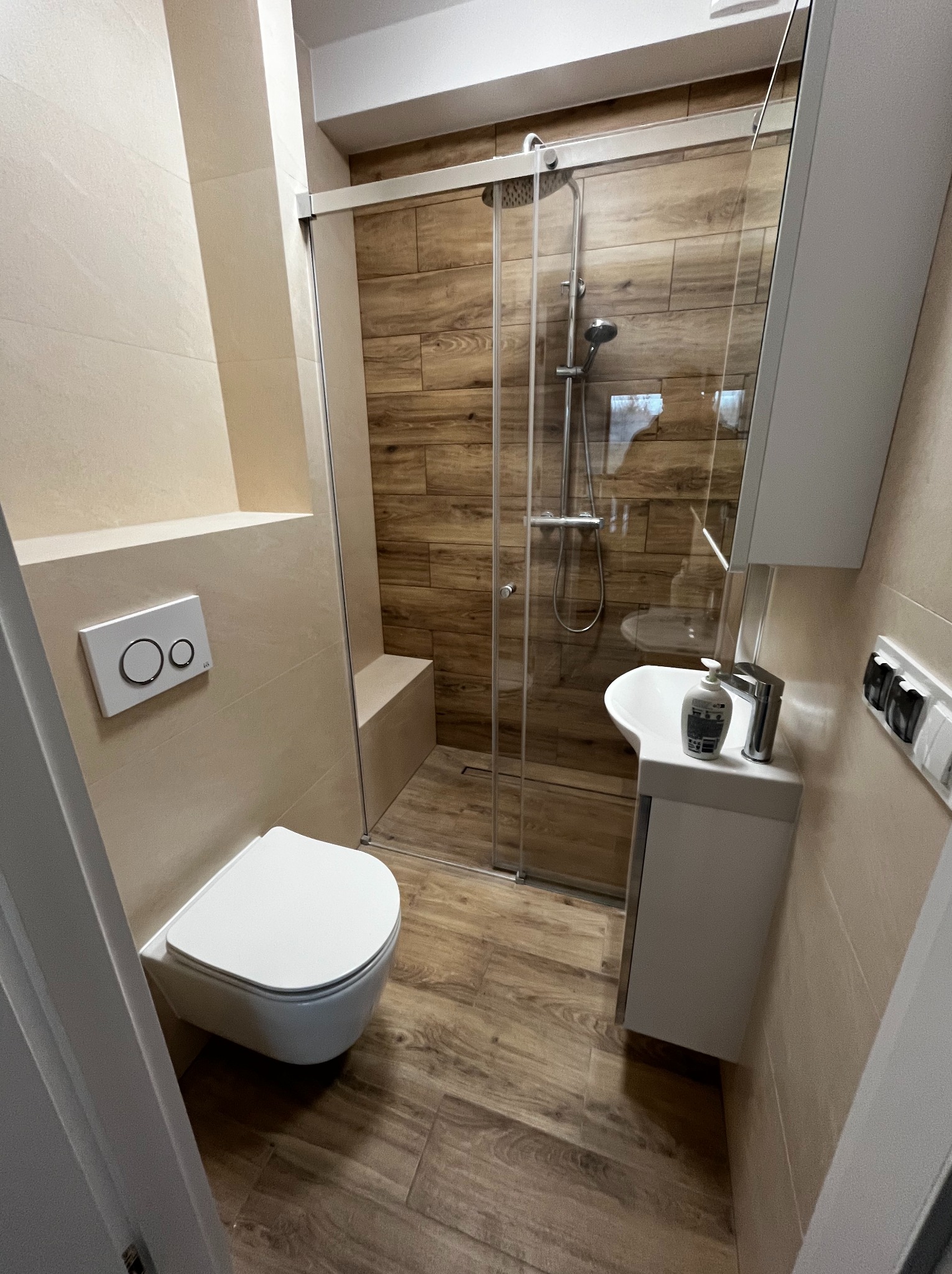 Nowoczesna łazienka z kabiną prysznicową typu walk-in, drewnopodobnymi płytkami i podwieszaną toaletą. Minimalistyczna umywalka z szafką i chromowaną armaturą.