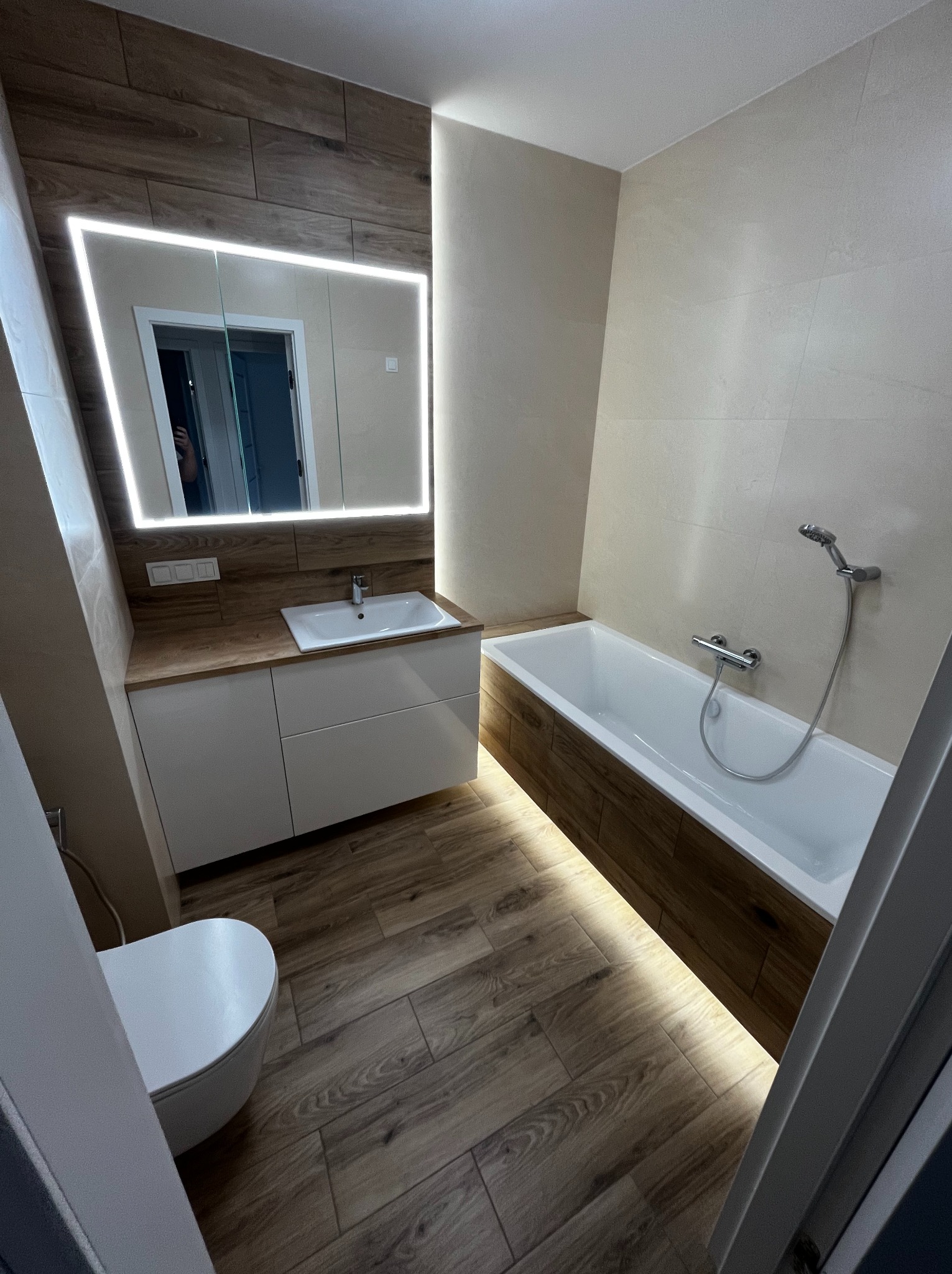 Nowoczesna łazienka z podświetlanym lustrem, drewnopodobnymi płytkami i białą armaturą. Uwagę zwraca podświetlenie wanny i szafki umywalkowej.