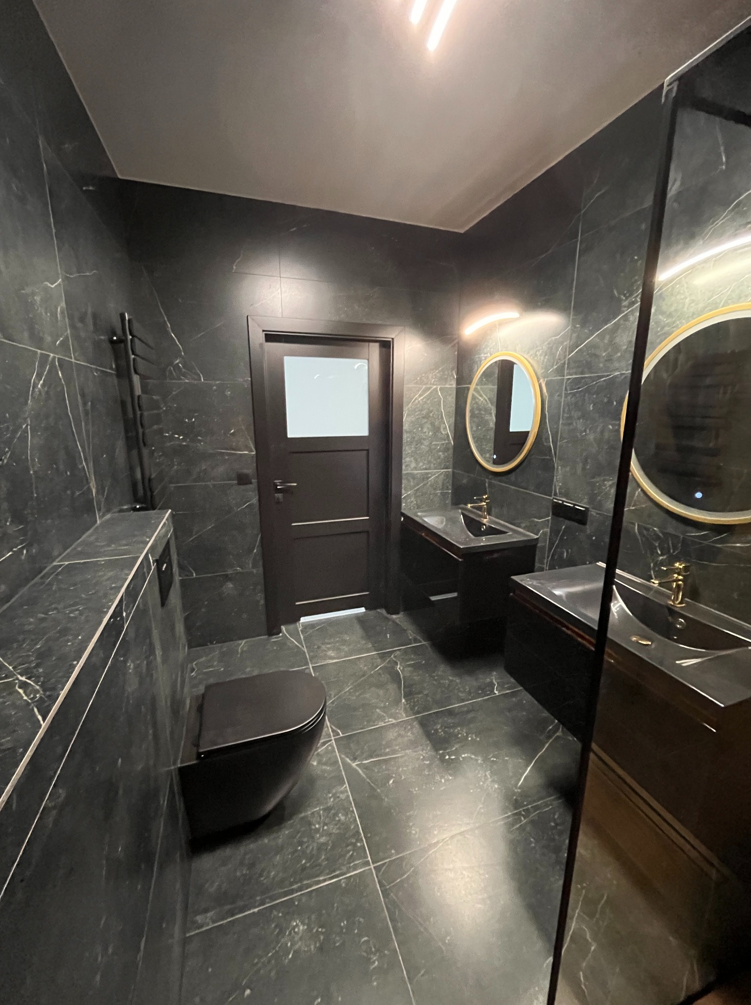 Elegancka, ciemna łazienka z marmurowymi płytkami, okrągłymi lustrami z podświetleniem i czarną armaturą. Minimalistyczny design z akcentem na luksusowe wykończenie.