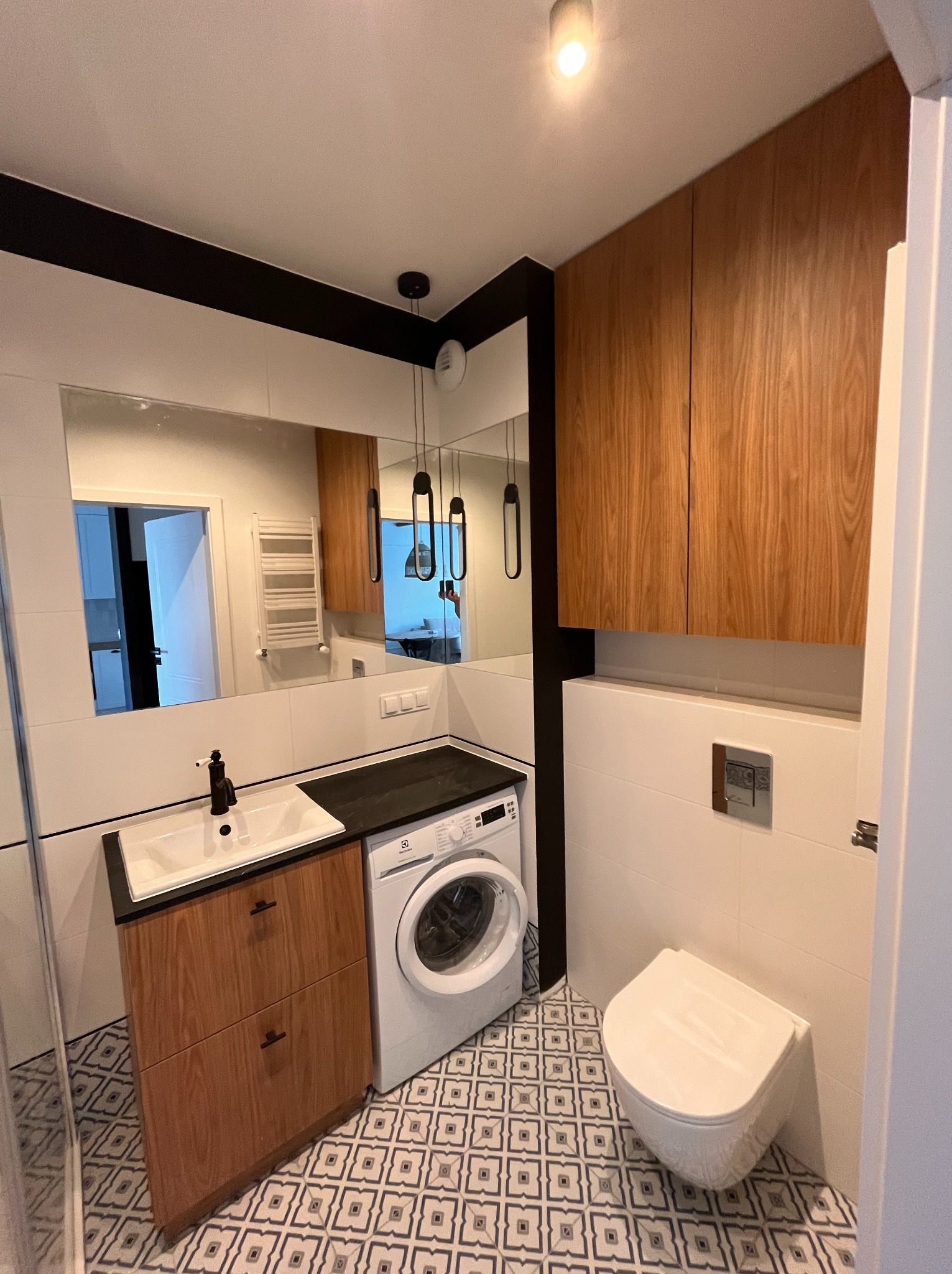 Nowoczesna łazienka z drewnianymi szafkami, umywalką z czarną armaturą, pralką i podwieszaną toaletą. Uwagę przykuwają wzorzyste płytki podłogowe.