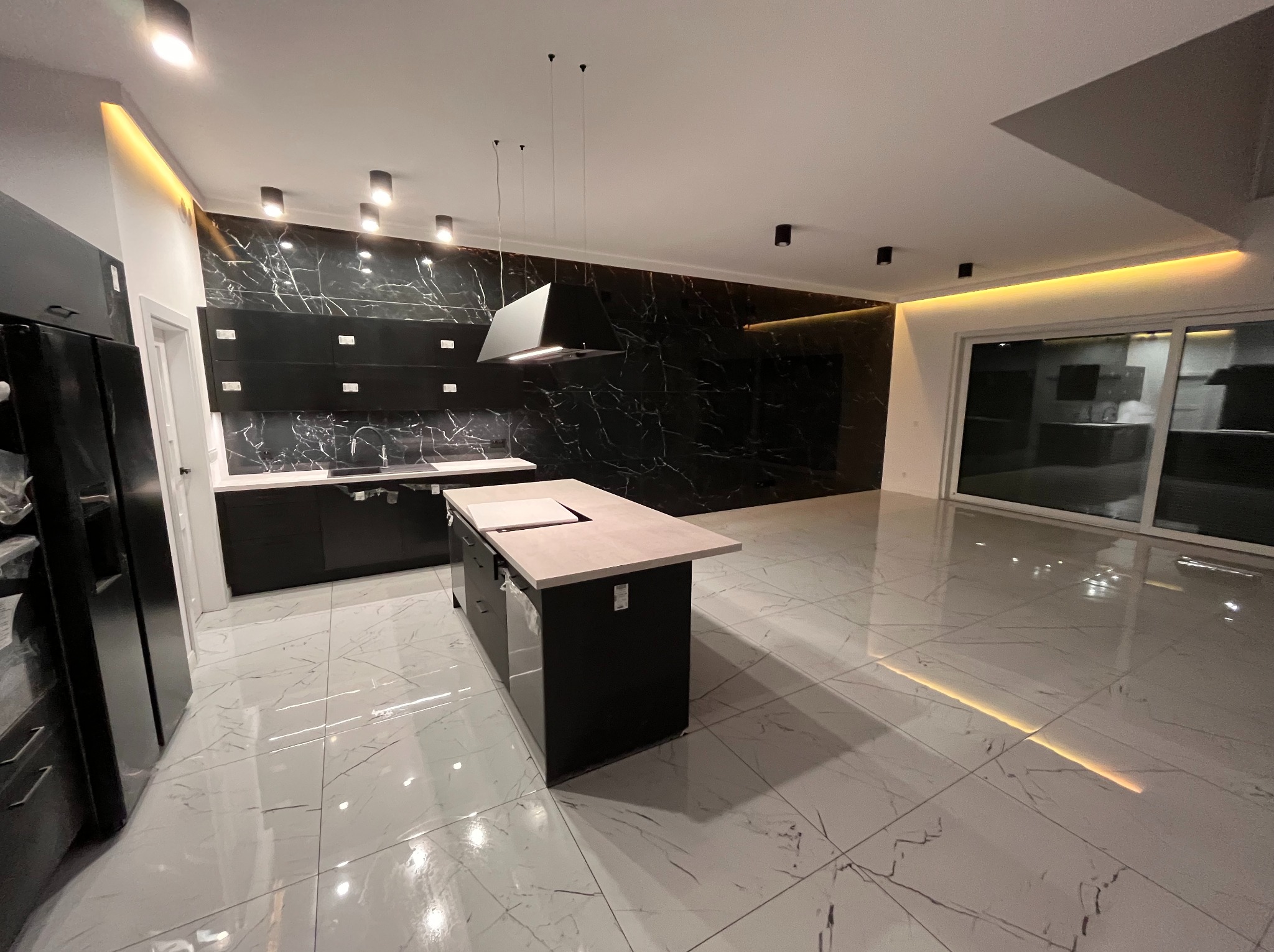 Nowoczesna kuchnia z wyspą, czarne szafki z marmurowym wzorem, biała podłoga z szarymi żyłami, oświetlenie LED. Minimalistyczny design, przestronne wnętrze.