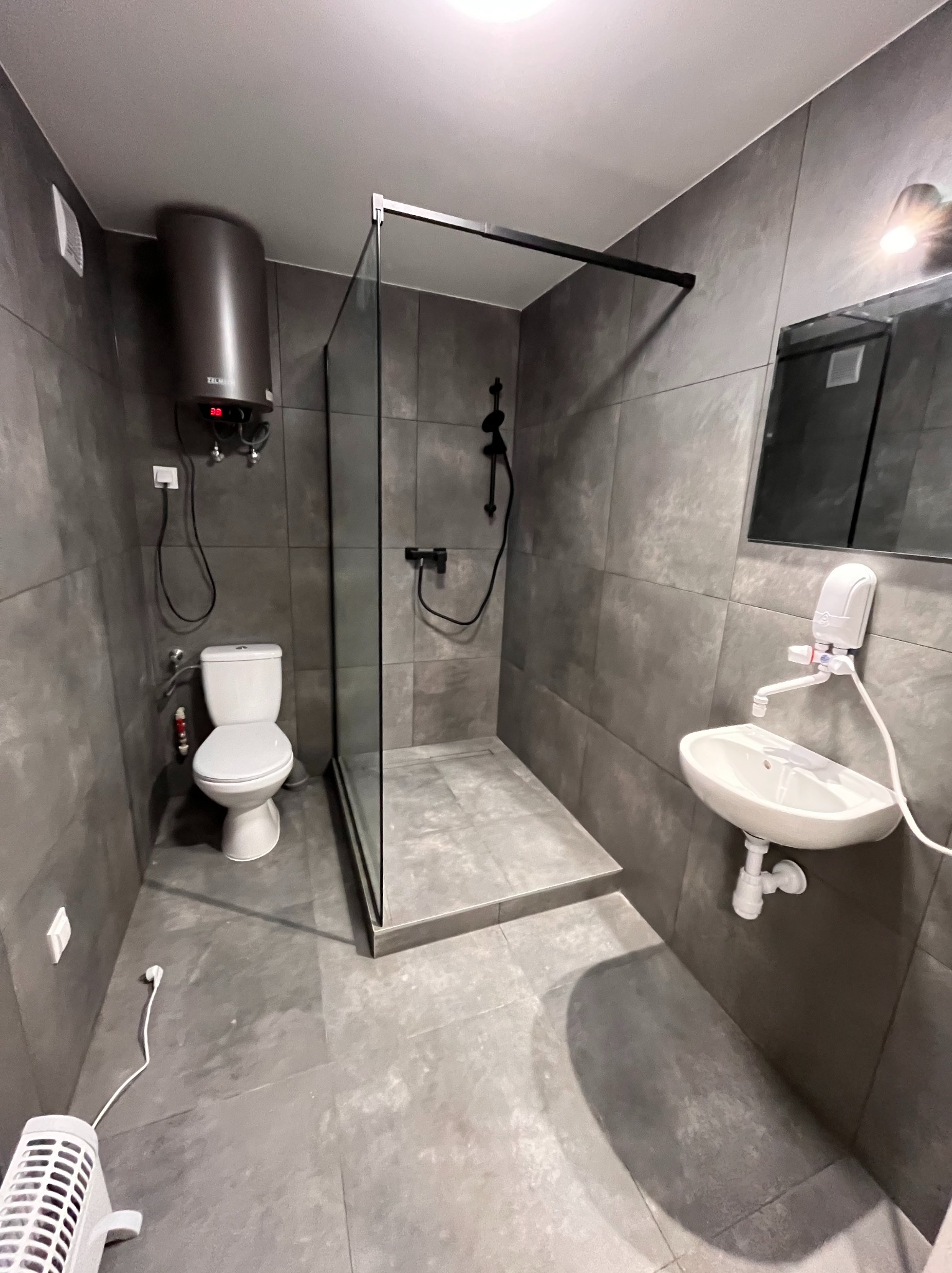 Nowoczesna łazienka w szarościach: kabina prysznicowa typu walk-in, podwieszana toaleta, umywalka nablatowa i bojler. Minimalistyczny design z akcentami czerni.