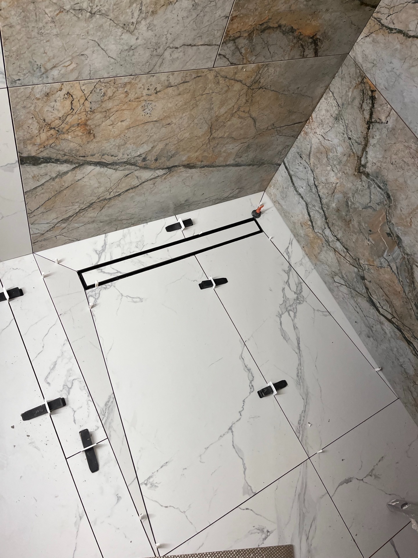 Ułożone płytki podłogowe i ścienne z marmurowym wzorem, czarny odpływ liniowy, czarne klipsy poziomujące, widok z góry, jasne oświetlenie.